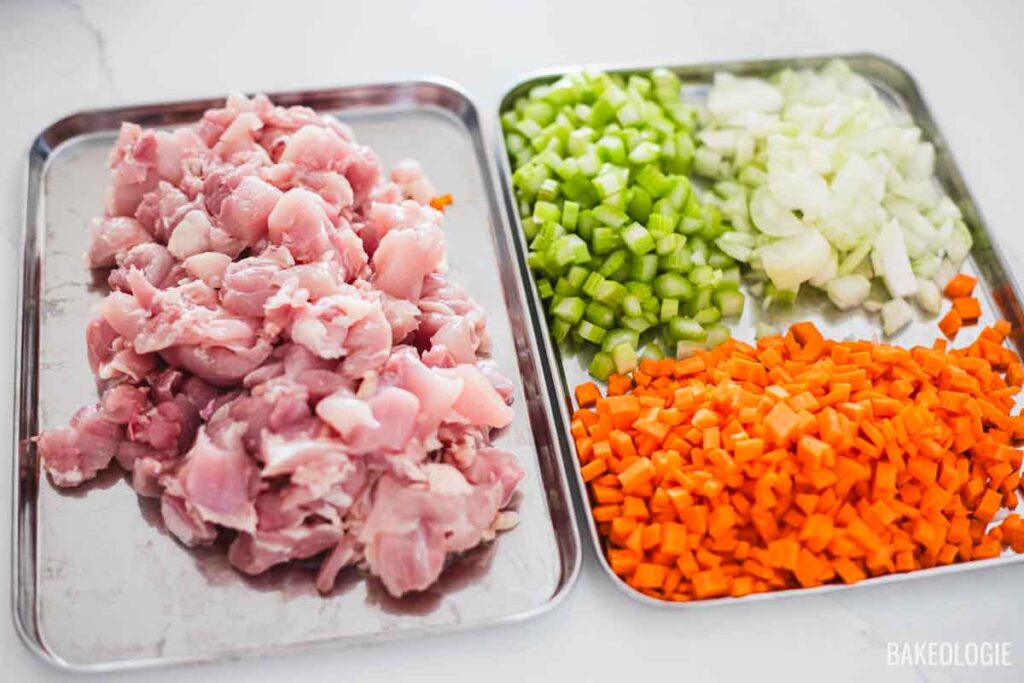 Chopped raw chicken, celery, onions, and carrots arranged on trays, prepped and ready for making chicken pot pie filling.