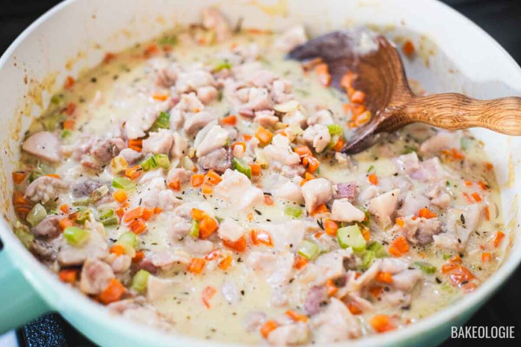 Creamy chicken pot pie filling simmering in a skillet with vegetables, bacon, and thyme before baking.
