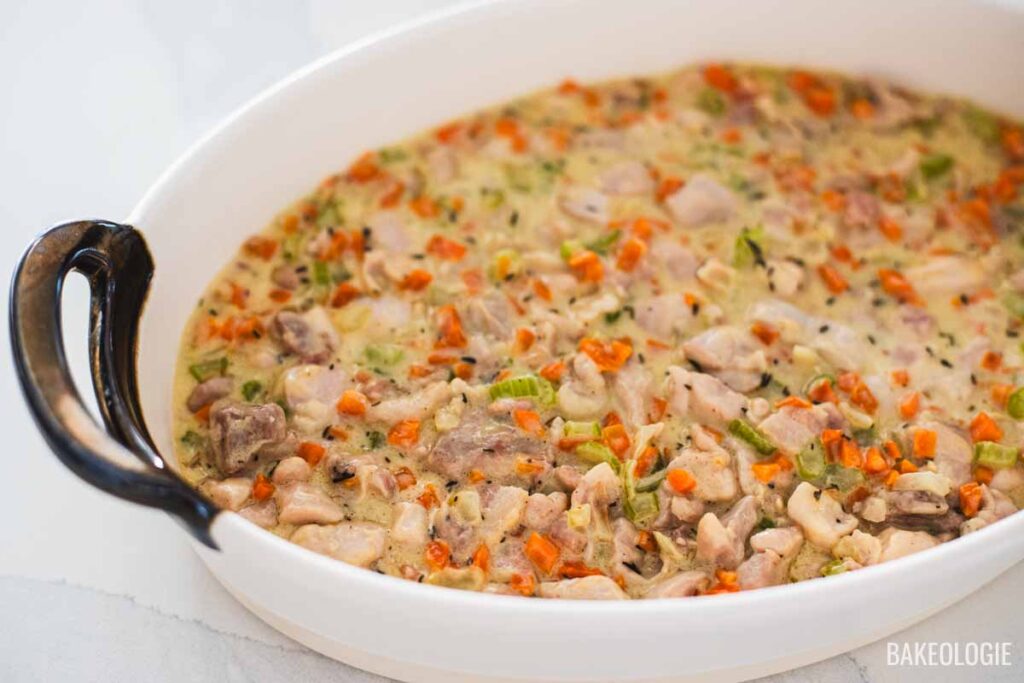 Creamy chicken pot pie filling with carrots, celery, and chicken spread evenly in a white oval baking dish before adding the puff pastry topping.