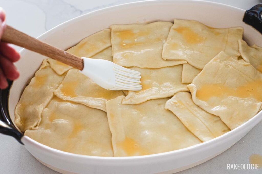 Brushing egg wash over puff pastry squares layered on top of chicken pot pie filling before baking to a golden finish.