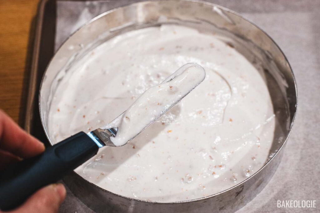 A hand using an offset spatula to spread cashew meringue evenly inside an 8-inch metal cake ring.