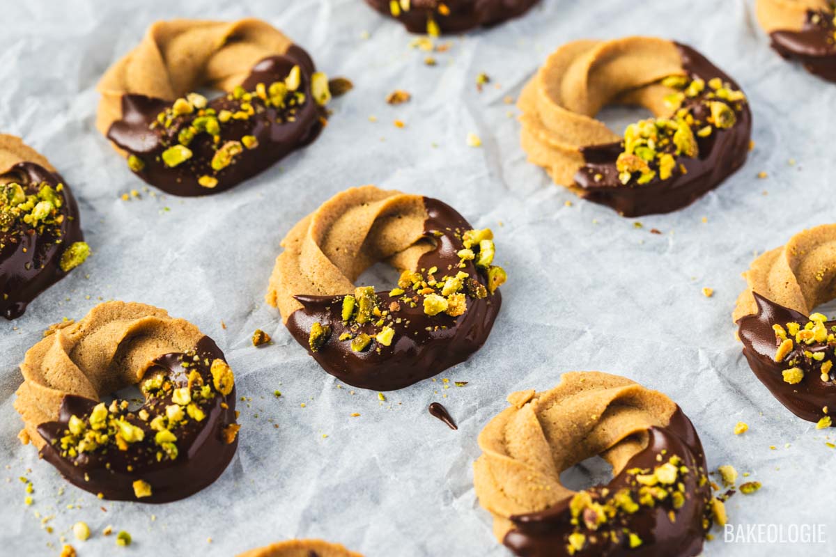 Coffee butter cookies with a piped swirl shape, dipped in chocolate and topped with chopped pistachios, cooling on parchment.
