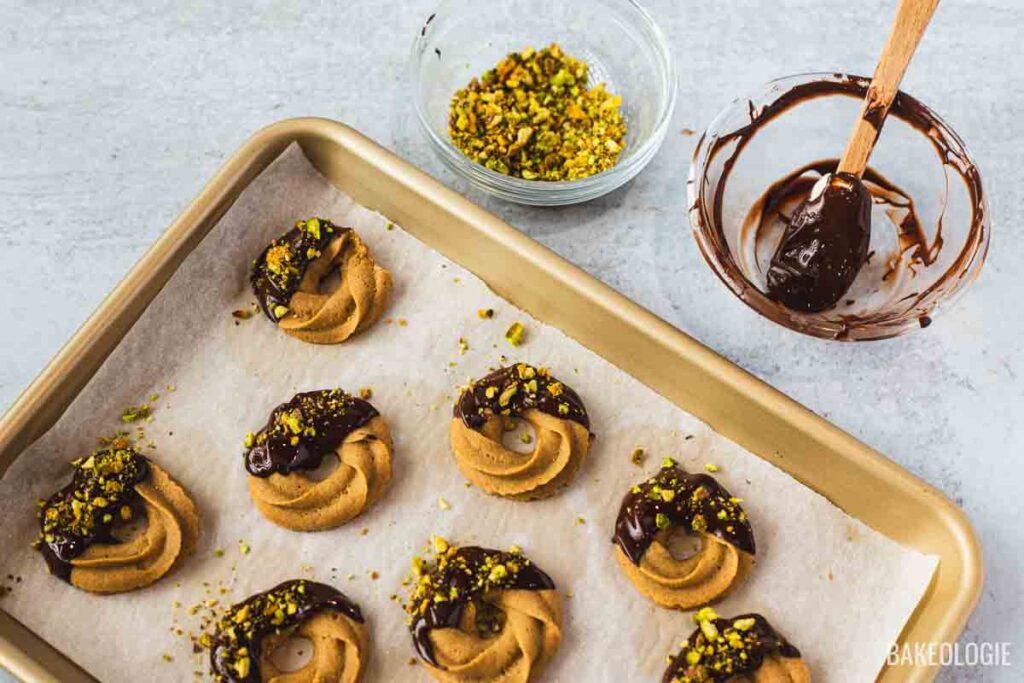 Baked coffee swirl cookies being dipped in melted chocolate and topped with chopped pistachios on a parchment-lined tray.