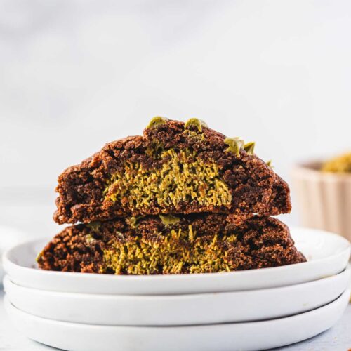 Inside view of a Dubai Chocolate Cookie cut in half, showing a thick gooey center filled with vibrant green pistachio chocolate. The cookie has a rich, crackly chocolate exterior and a soft, fudgy pistachio middle, stacked on white plates with crumbs scattered around.