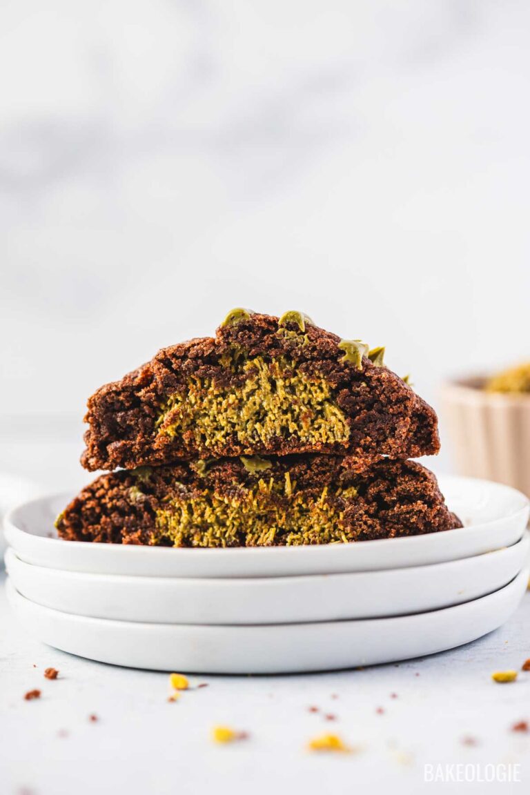 Inside view of a Dubai Chocolate Cookie cut in half, showing a thick gooey center filled with vibrant green pistachio chocolate. The cookie has a rich, crackly chocolate exterior and a soft, fudgy pistachio middle, stacked on white plates with crumbs scattered around.