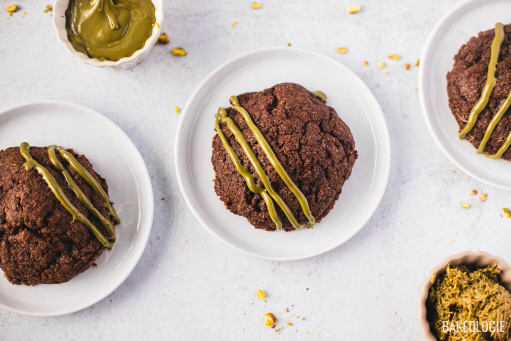Three Dubai Chocolate Cookies on white plates, each with a crackly chocolate top and drizzled with glossy pistachio chocolate. Small bowls of pistachio filling and chopped pistachios sit nearby, with crumbs scattered on a light background.
