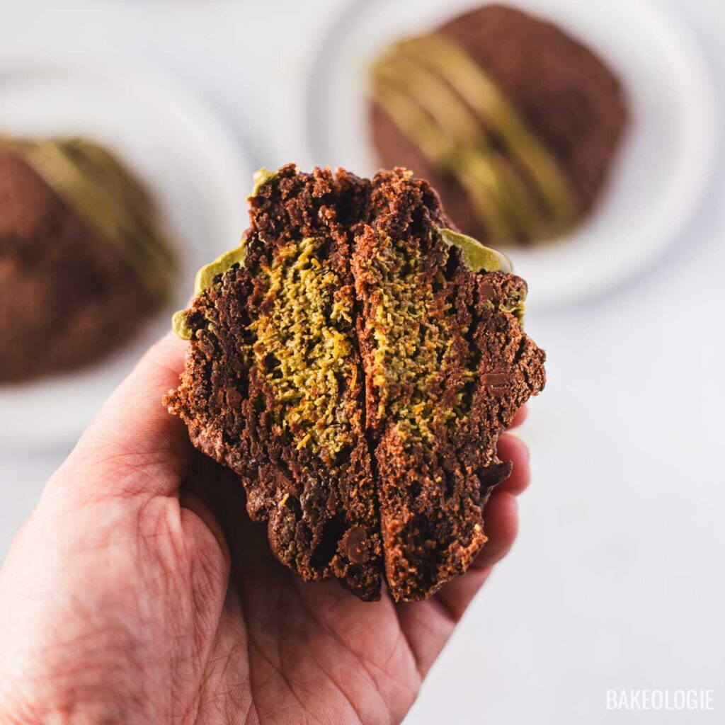 Three Dubai Chocolate Cookies on white plates, each with a crackly chocolate top and drizzled with glossy pistachio chocolate. Small bowls of pistachio filling and chopped pistachios sit nearby, with crumbs scattered on a light background.