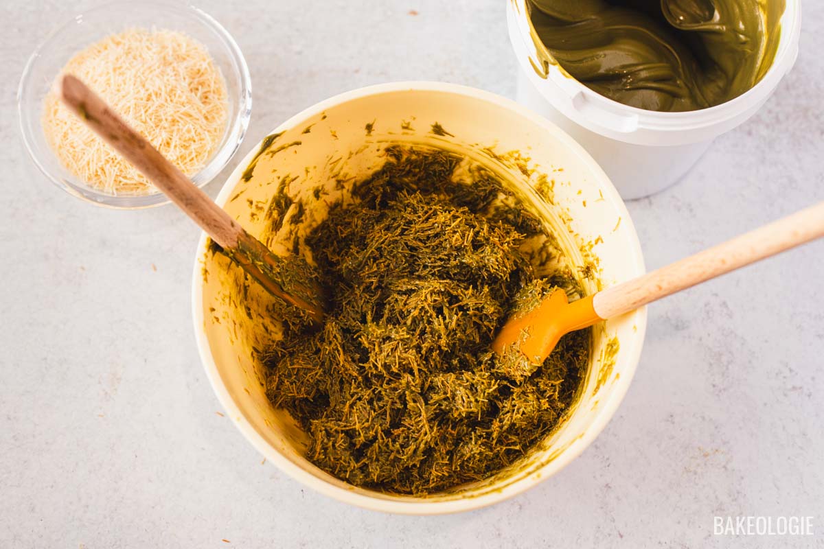 Bowl of toasted kataifi mixed with pistachio filling, showing the green, shredded mixture being stirred with wooden and silicone spatulas. A bowl of plain toasted kataifi sits beside it on a light surface.