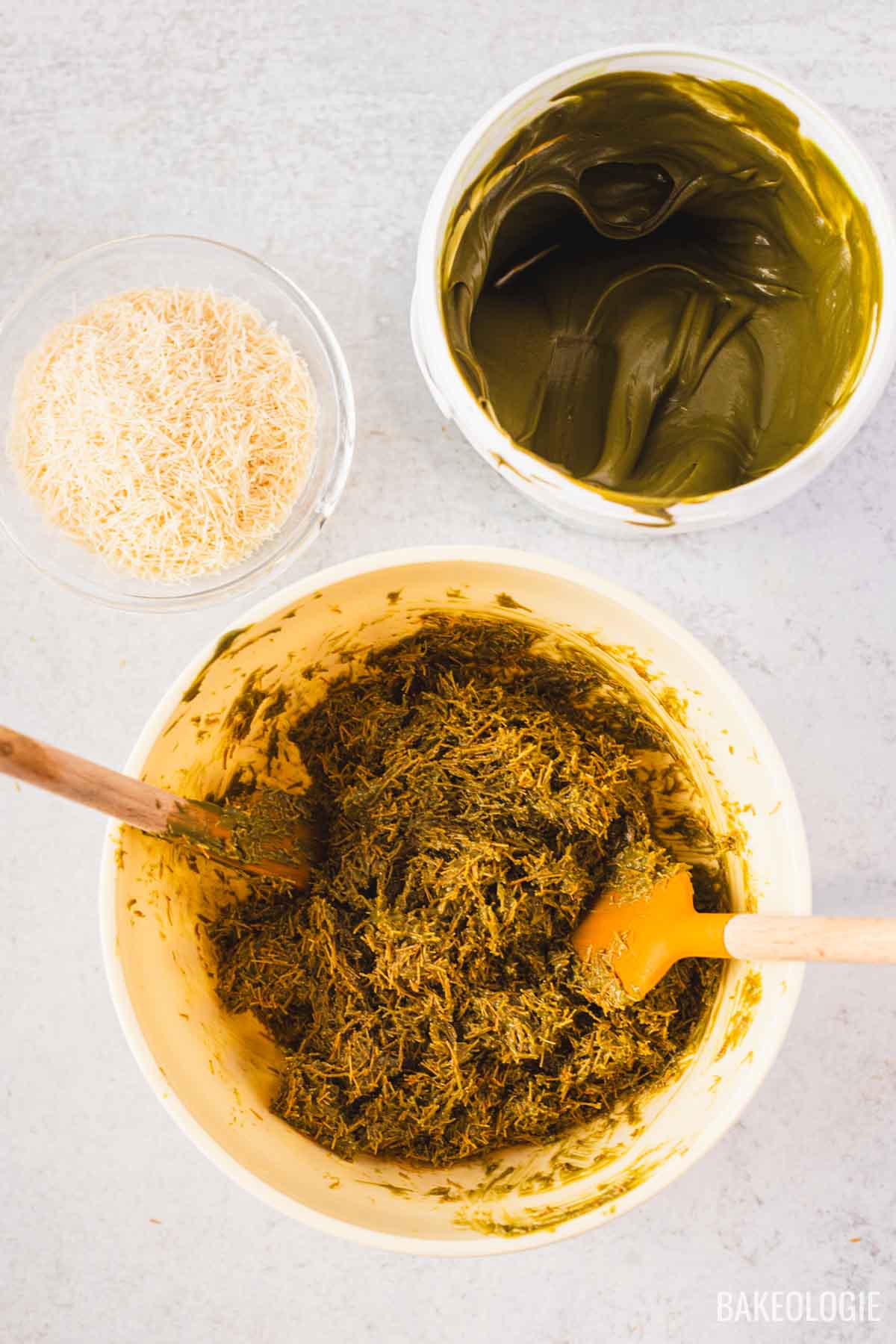Top-down view of pistachio kataifi being mixed in a large bowl, with a container of smooth pistachio filling and a bowl of toasted kataifi strands arranged beside it.