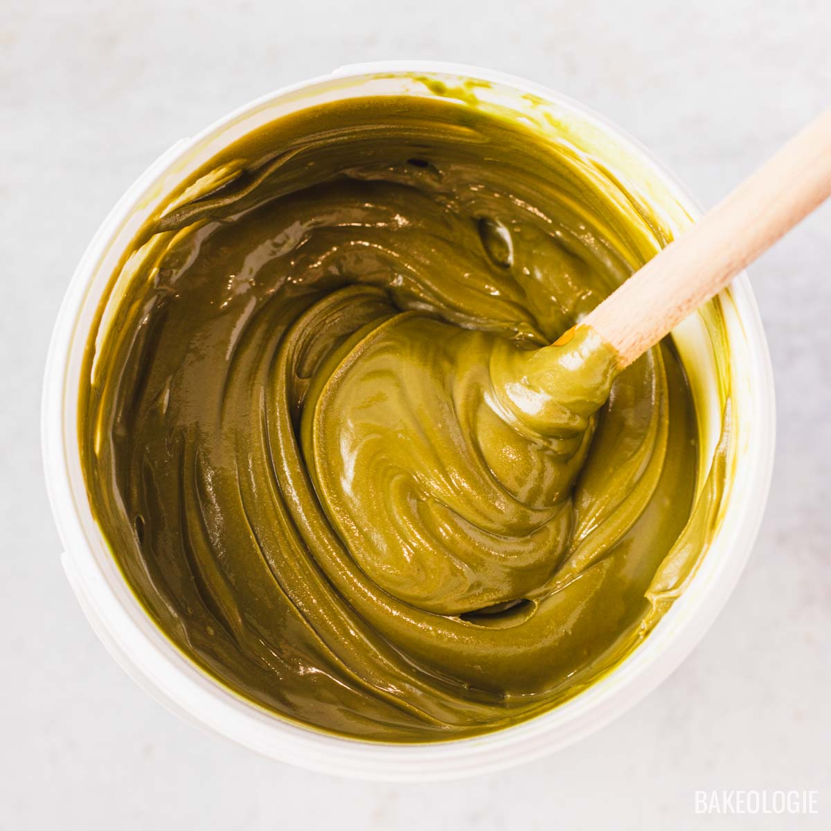 Close-up of a bucket filled with glossy green pistachio filling, with a wooden spatula dipped into the smooth mixture.