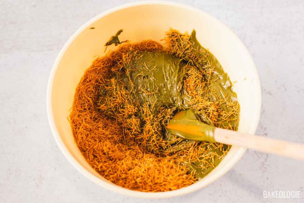 Toasted kataifi being mixed into smooth pistachio cream in a large bowl, with a spatula folding the strands into the mixture.