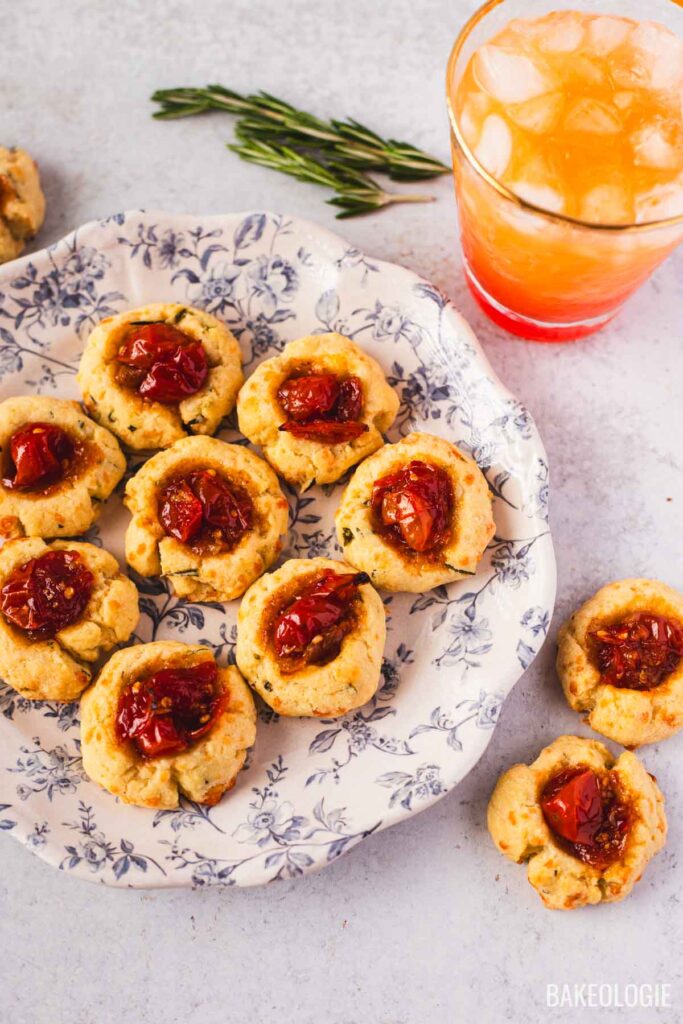 Herbed cheese thumbprint cookies with tomato jam centers arranged on a floral plate, served as a savory appetizer with a cocktail.