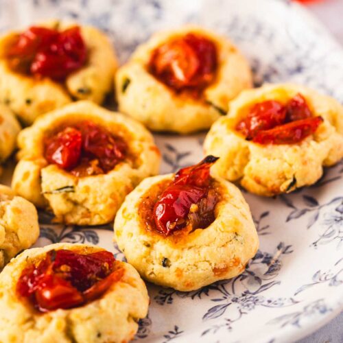 Close-up of herbed cheese thumbprint cookies filled with tomato jam, showing the crumbly cheese texture and glossy jam topping.