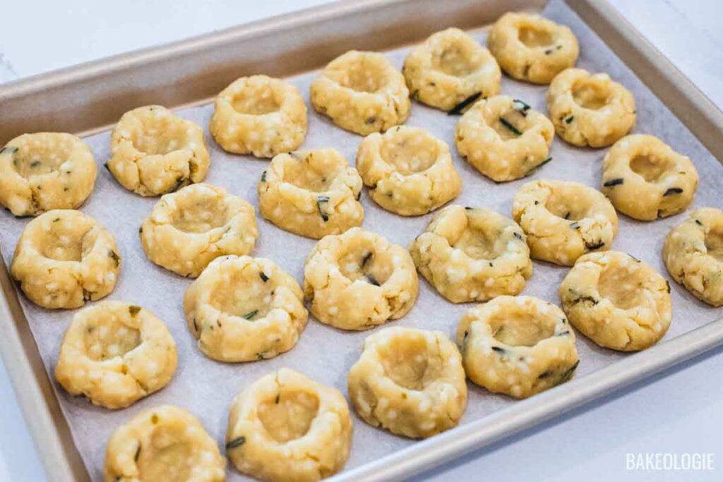 Unbaked herbed cheese thumbprint cookies shaped and pressed with deep wells, arranged on a parchment-lined baking tray ready for freezing.