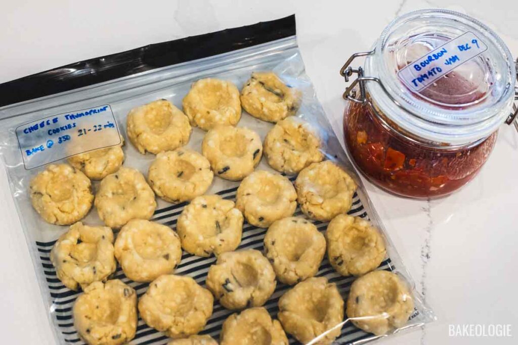 Shaped herbed cheese thumbprint cookies stored flat in a freezer bag alongside a jar of tomato jam for make-ahead baking.