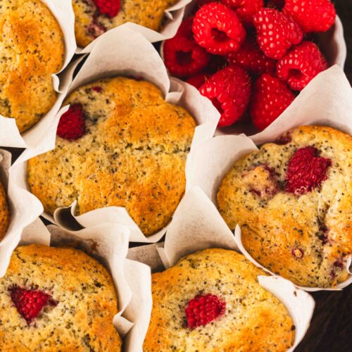 Close-up of bakery-style raspberry lemon poppyseed muffins baked in tulip parchment liners, featuring golden domed tops with fresh raspberries and poppy seeds, styled beside a bowl of fresh raspberries.