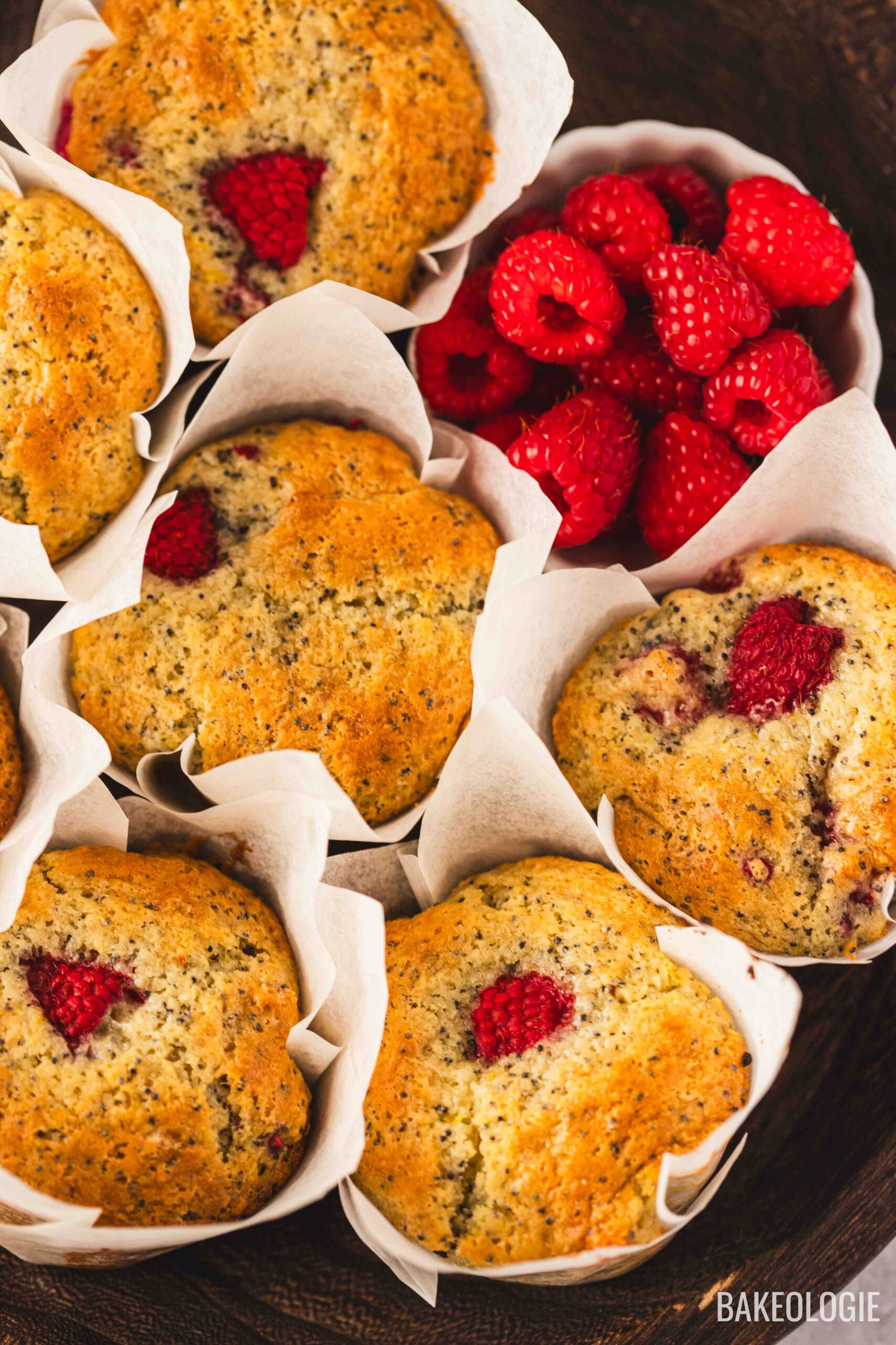 Close-up of bakery-style raspberry lemon poppyseed muffins baked in tulip parchment liners, featuring golden domed tops with fresh raspberries and poppy seeds, styled beside a bowl of fresh raspberries.