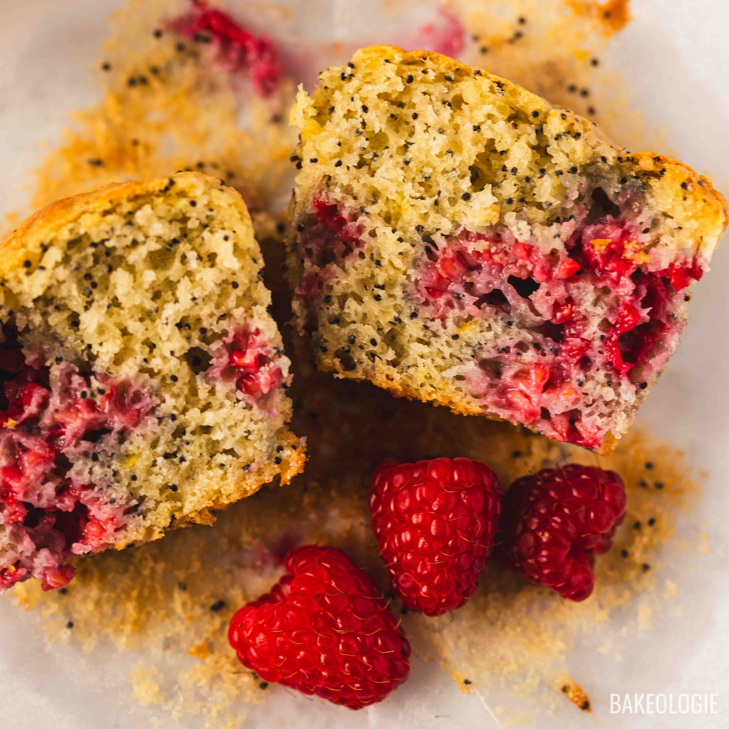 Close-up of a raspberry lemon poppyseed muffin cut in half, showing a soft, moist crumb with poppy seeds and a juicy raspberry center, styled with fresh raspberries on parchment paper.