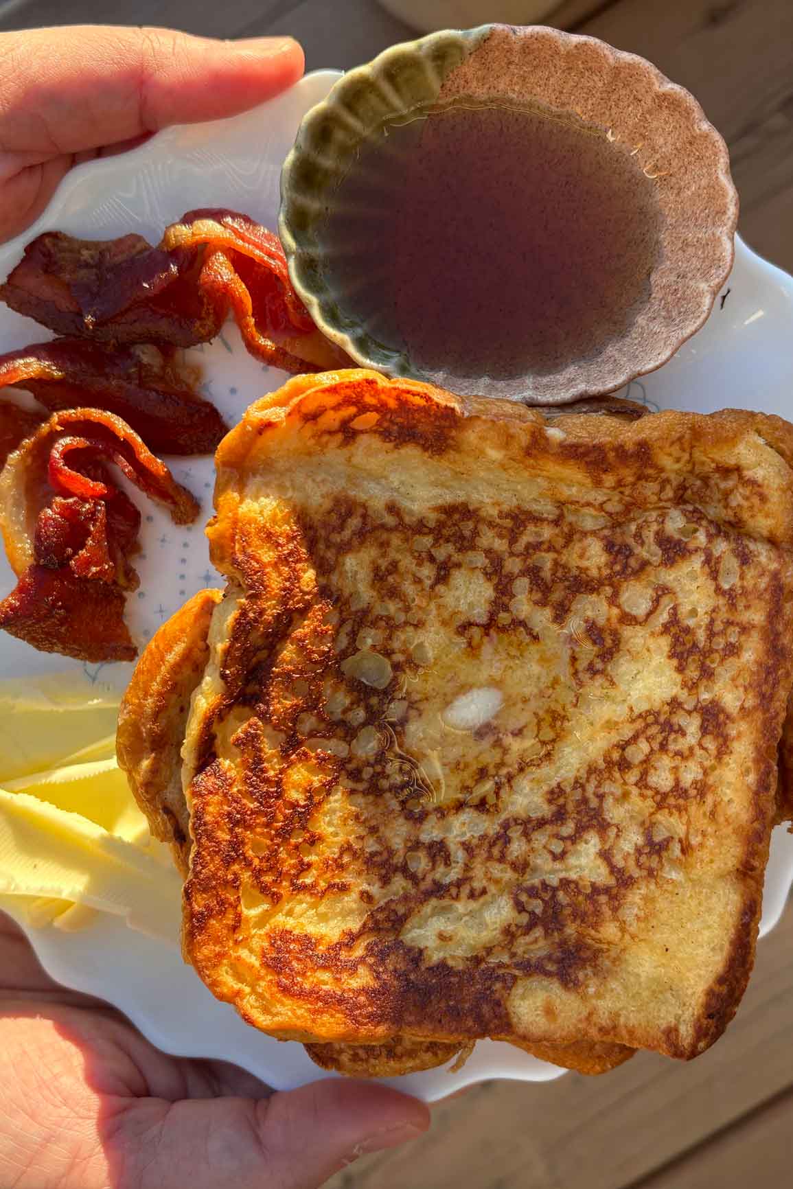 Golden brown cinnamon French toast with crisp edges, served with butter, crispy bacon, and maple syrup in a small bowl.