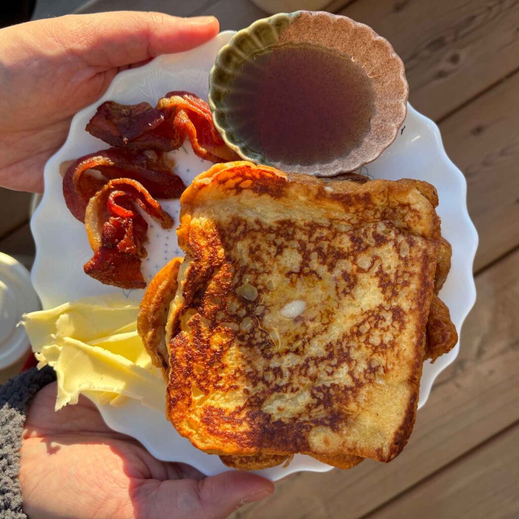 Golden brown cinnamon French toast with crisp edges, served with butter, crispy bacon, and maple syrup in a small bowl.