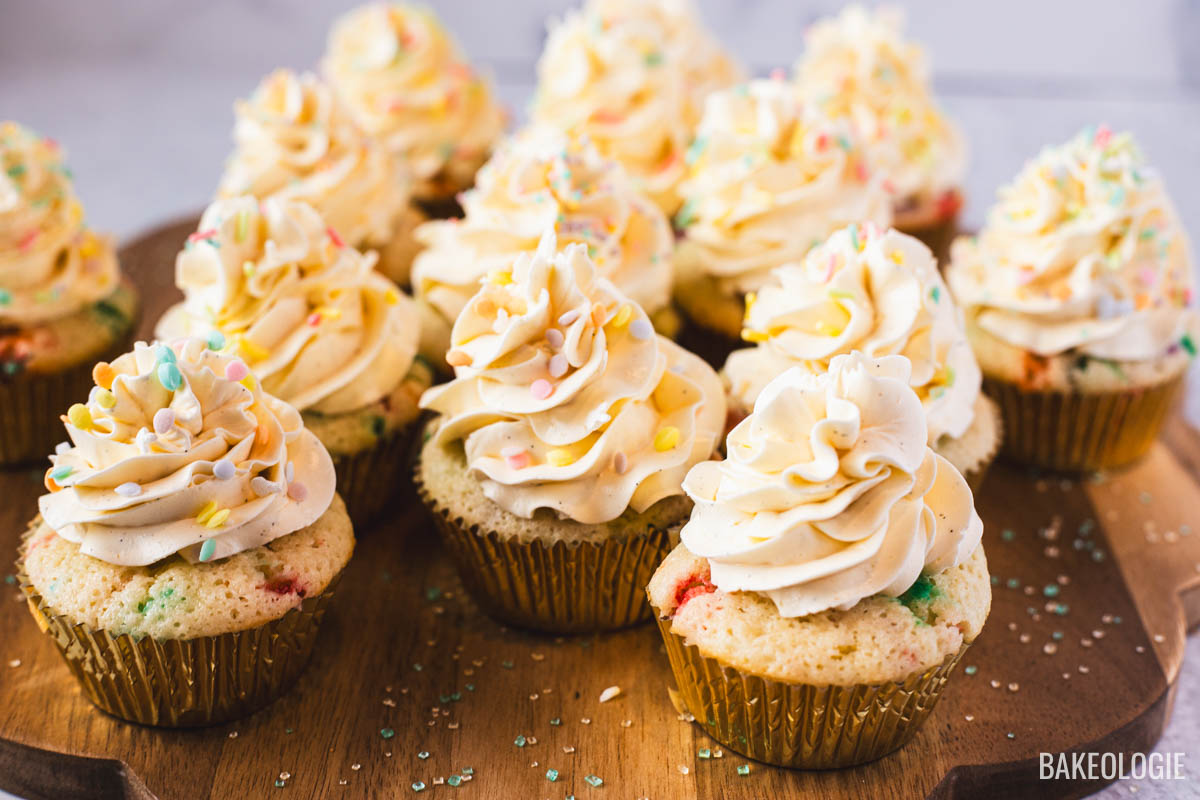 Vanilla cupcakes topped with piped Swiss meringue buttercream and colorful sprinkles on a wooden board