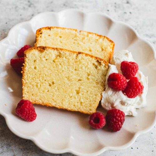 Two slices of cream cheese pound cake on a scalloped ceramic plate, served with a dollop of whipped cream and fresh raspberries. The cake has a golden crust and a moist, tender crumb with a fine texture.