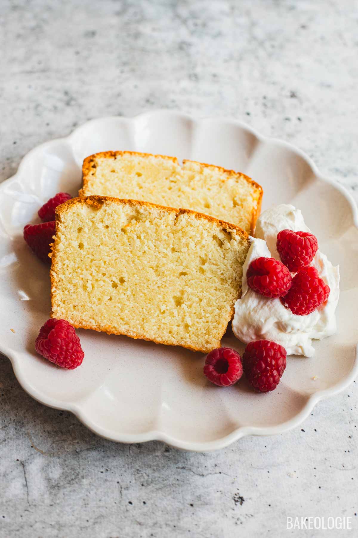 Two slices of cream cheese pound cake on a scalloped ceramic plate, served with a dollop of whipped cream and fresh raspberries. The cake has a golden crust and a moist, tender crumb with a fine texture.