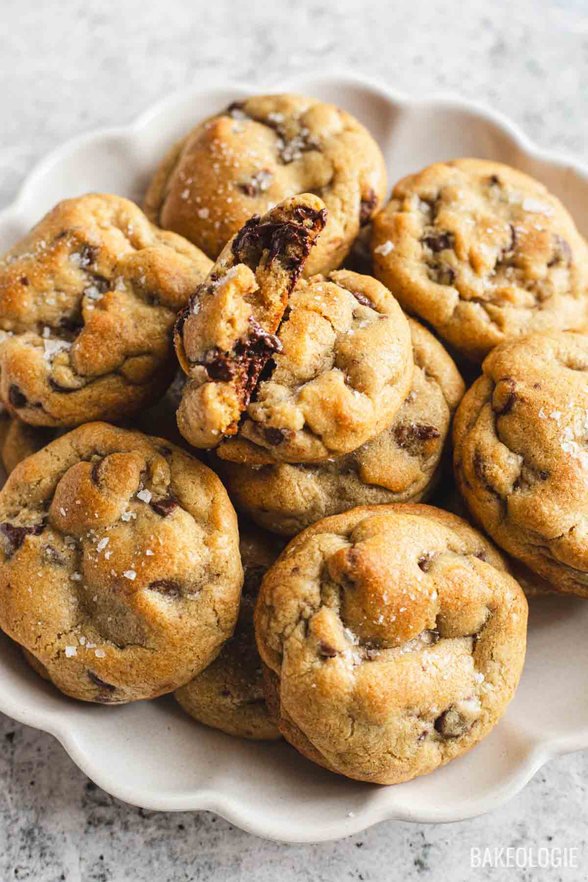 Stack of chewy chocolate chip cookies with flaky sea salt and melted chocolate chunks on a white serving plate.