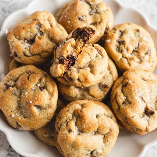 Stack of chewy chocolate chip cookies with flaky sea salt and melted chocolate chunks on a white serving plate.