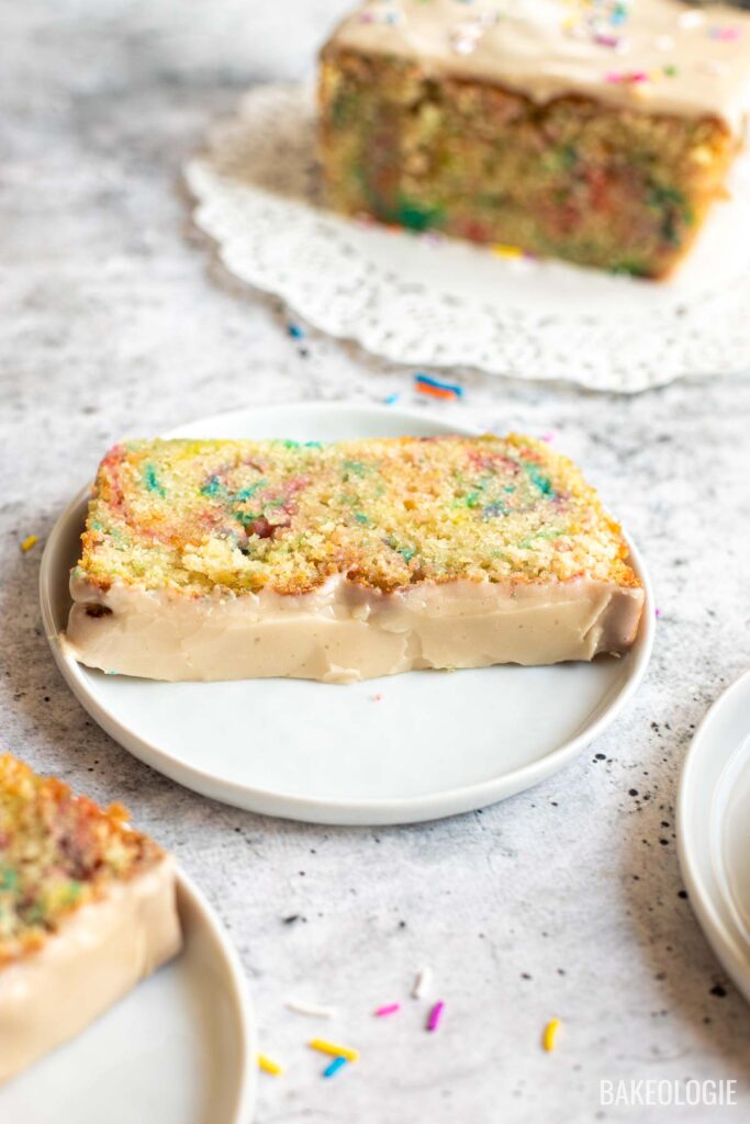 Slice of Funfetti Sour Cream Pound Cake with vanilla glaze served on a white plate with the loaf in the background.