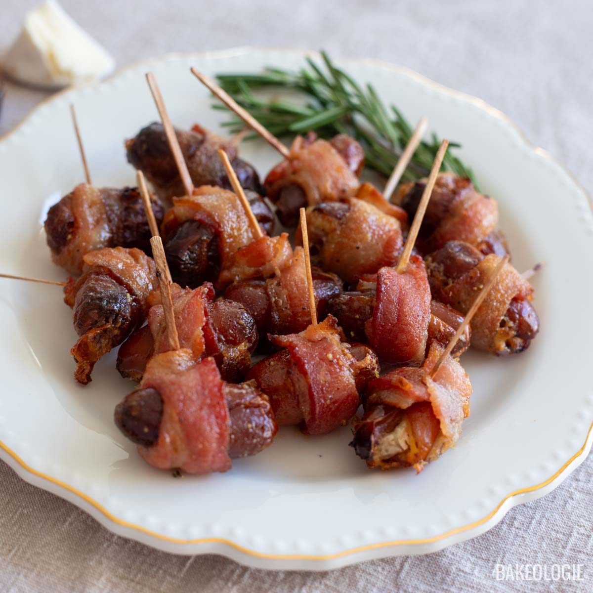 bacon wrapped dates with maple glaze on a white platter with rosemary sprigs and slice of cheese on the side