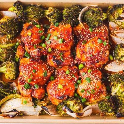 Honey gochujang chicken thighs baked with roasted broccoli and onions, topped with sesame seeds and sliced green onions.